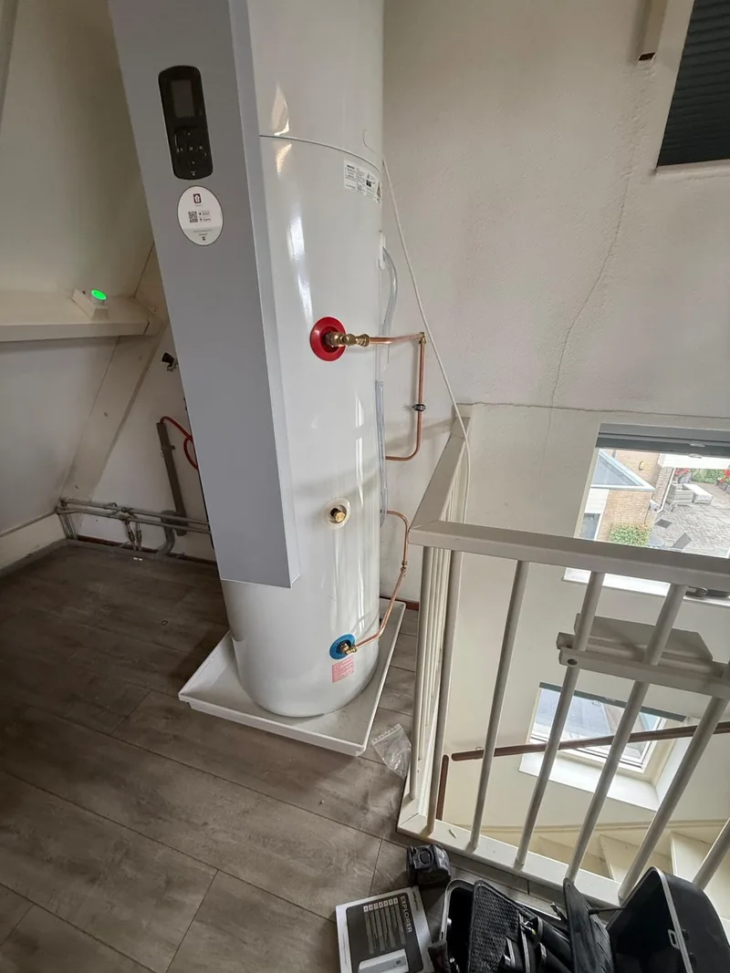 Warmtepompboiler op zolder met verchroomd koperen leidingwerk en lekbak, naast het trapgat