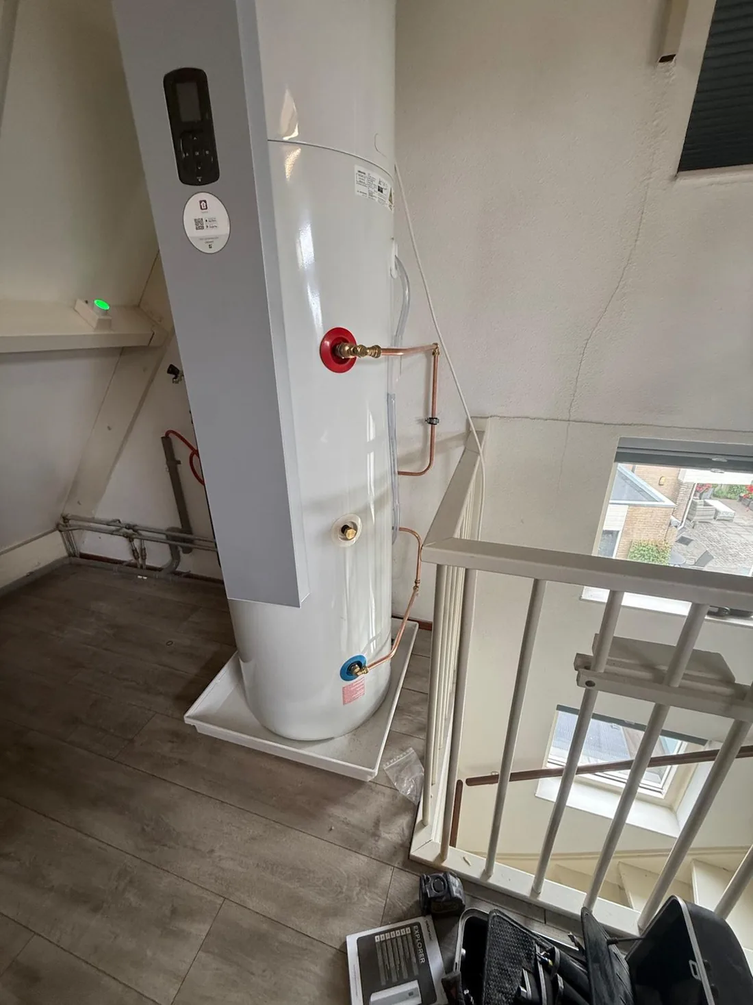 Warmtepompboiler op zolder met koperen aansluitingen en lekbak, naast het trapgat